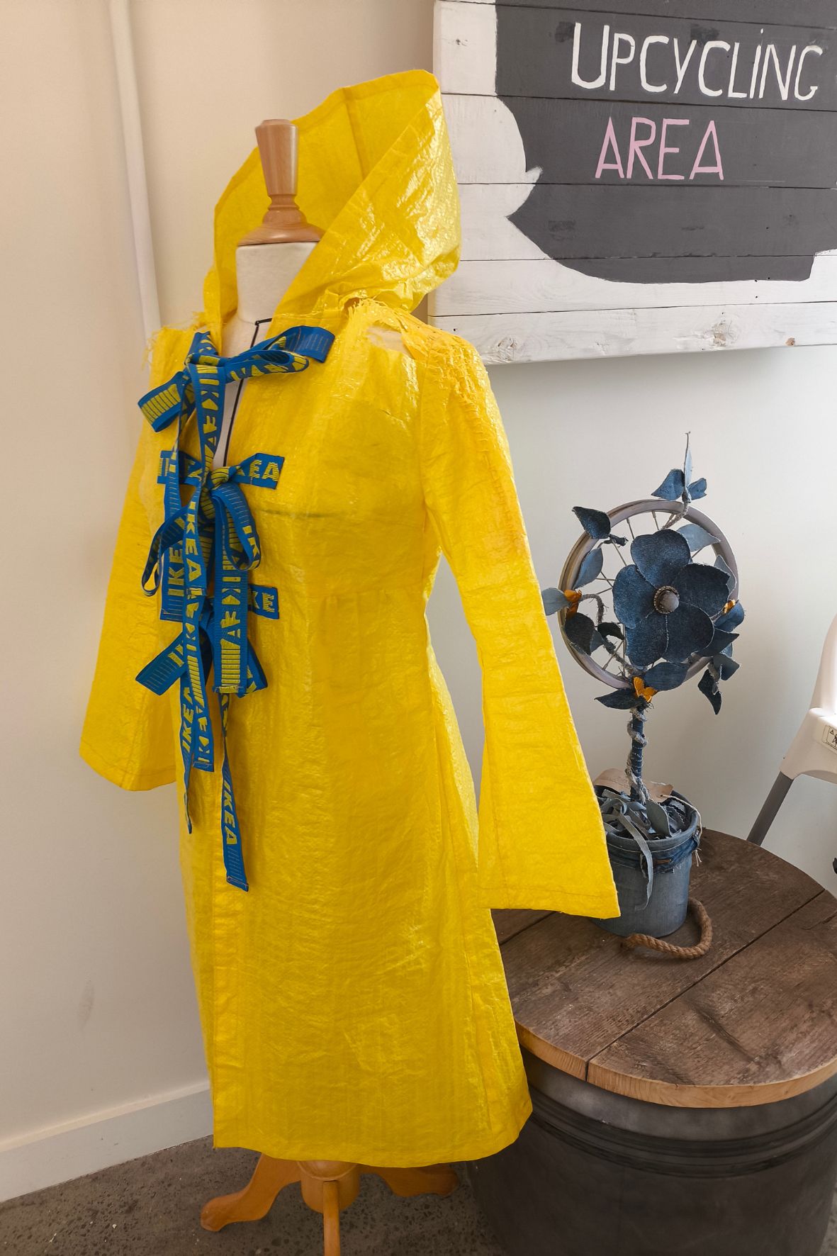 Bild zeigt einen gelben Regemantel, gemacht aus alten Ikea-Permanenttragetaschen. Dahinter ein Schild mit der Aufschrift: Upcycling Area.