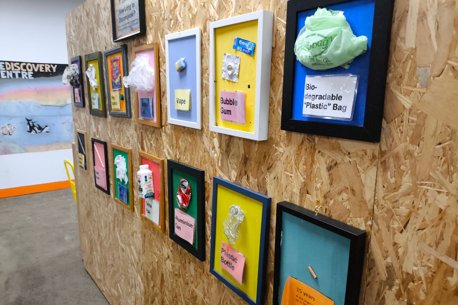 Bild zeigt Tafeln auf einer Holzwand mit unterschiedlichen Produkten und deren biologischen Abbaubarkeit. 